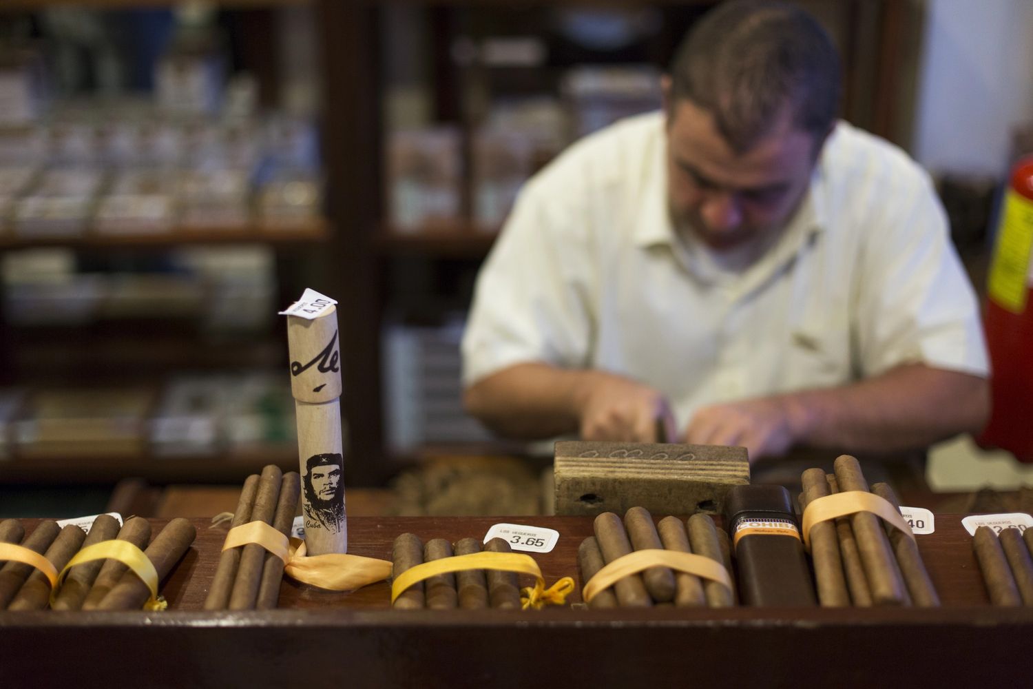 Handcrafted cigars with price tags on display at a Tulsa cigar store, with a craftsman working in the background.