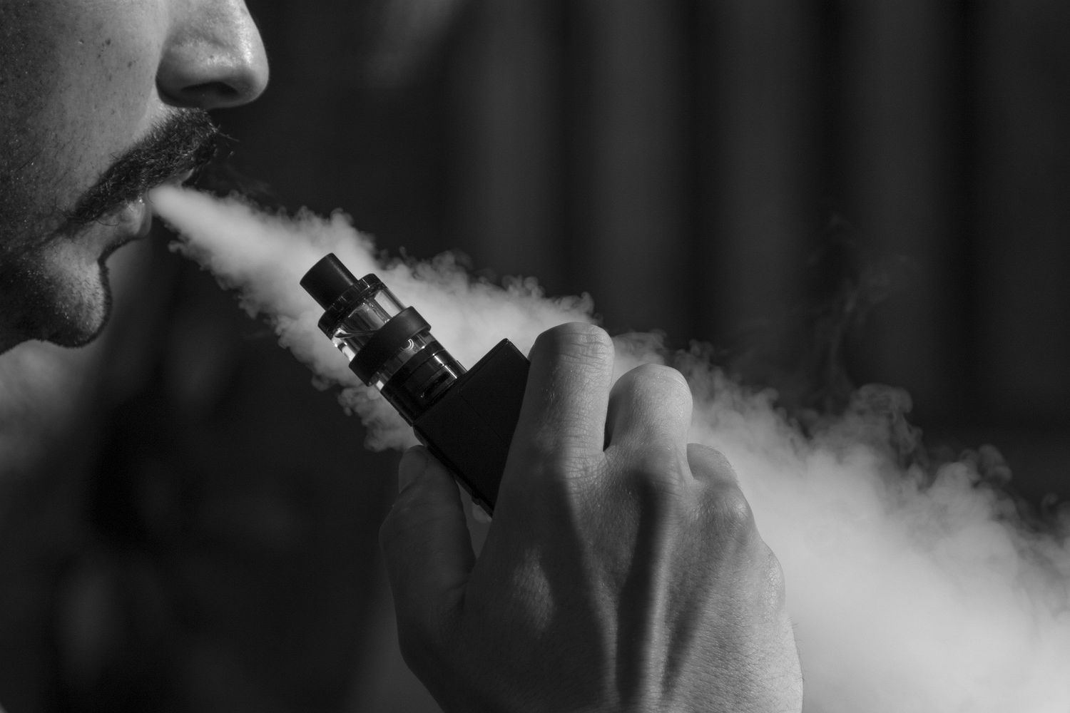 Black and white photo of a man exhaling a large cloud of vapor from a premium box Vape Mods Tulsa device.