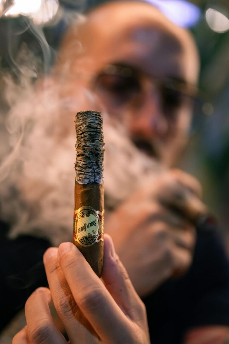 Close-up of a hand holding a well-lit Brick House cigar with ash at a comfortable Cigar Shop Tulsa.