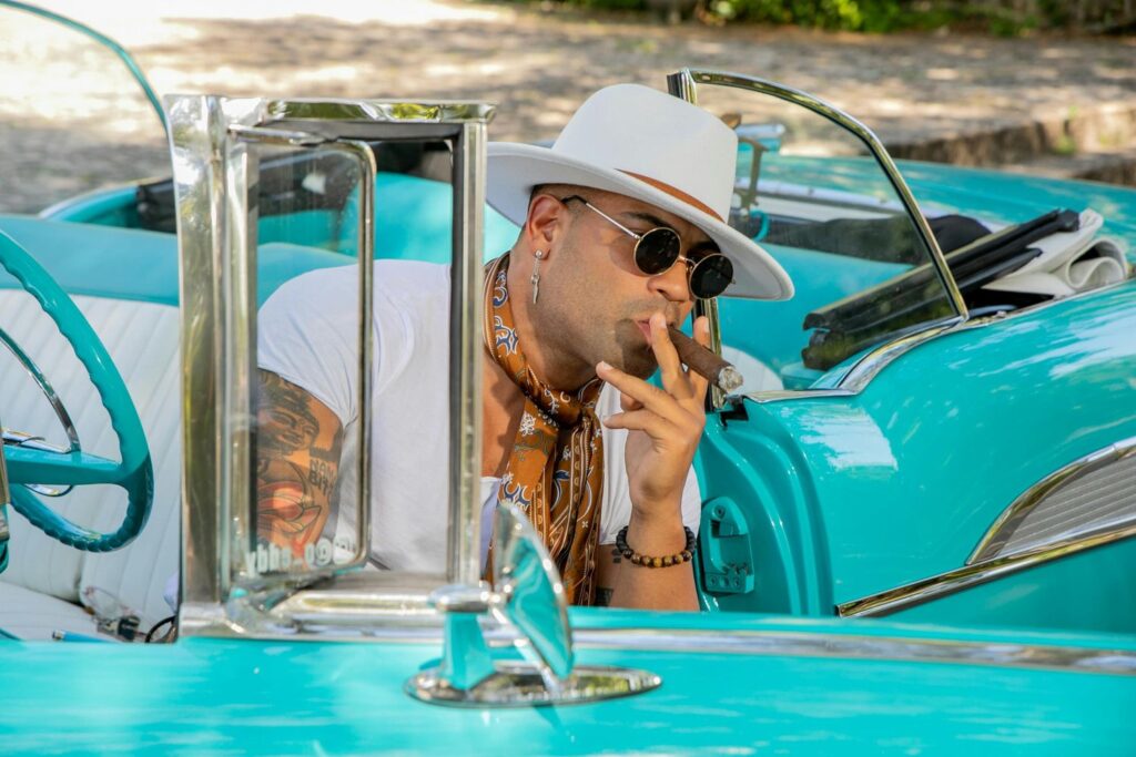 Stylish man in a white hat smoking a cigar in a vintage turquoise convertible, promoting Cigar Shop Tulsa.
