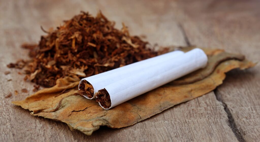 Two handmade cigarettes lying on a dried tobacco leaf with a pile of loose tobacco nearby. Find quality tobacco at Tobacco Shop Tulsa.