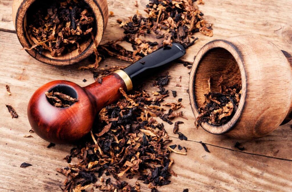 A wooden pipe surrounded by loose pipe tobacco and two wooden cups on a rustic table. Buy pipe tobacco at Tobacco Shop Tulsa.