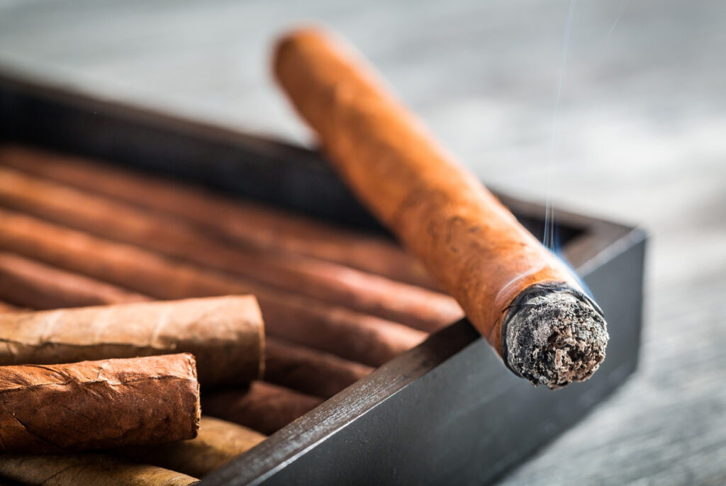 Close up of a burning cigar resting on a wooden humidor box filled with fresh tobacco sticks at a cigar store Tulsa.