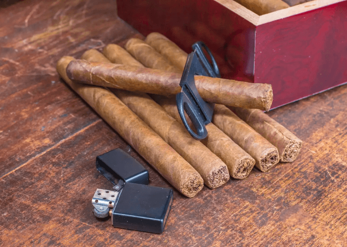 A row of cigars, a black flip lighter, and a cutter near a wooden humidor at a local cigar shop Tulsa destination.