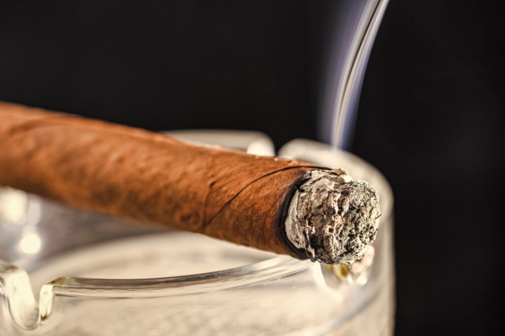A premium hand-rolled cigar with fine grey ash and wisps of smoke resting on an ashtray at a cigar shop Tulsa.