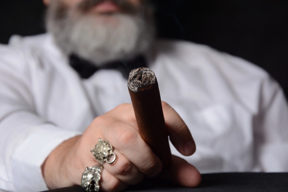 A man with a grey beard and black bowtie holds a smoking, lit tobacco roll, perfect for a cigar store tulsa blog.