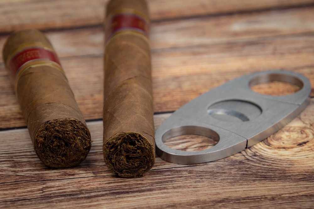 A pair of premium rolled tobacco sticks lie on a wooden table beside a metallic cutter from a cigar store tulsa shop.