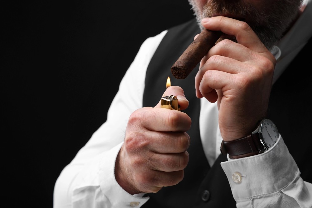 An close-up view of a man in a vest sparking a golden lighter to smoke a premium roll from a cigar store tulsa selection.