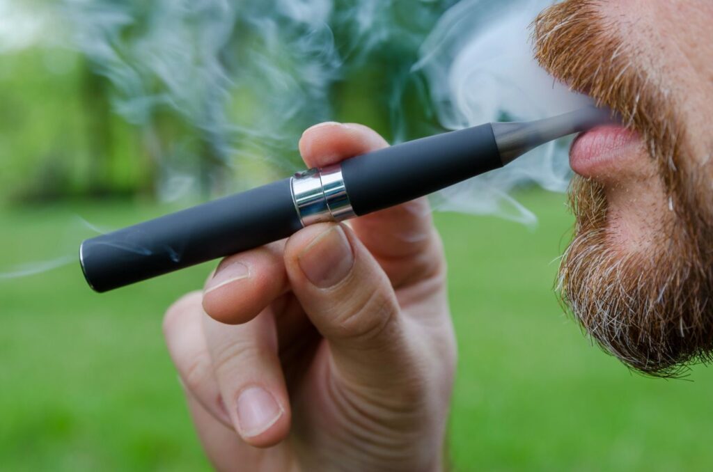 A bearded man enjoying e-cigarettes Tulsa outdoors, holding a sleek black vape pen while exhaling vapor in a park.
