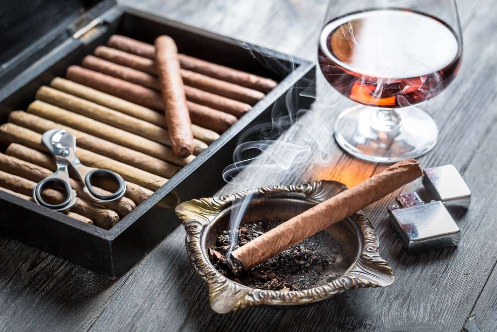 Luxury cigar shop Tulsa selection with a wooden humidor box, metal cutters, a smoking cigar, and a glass of brandy.