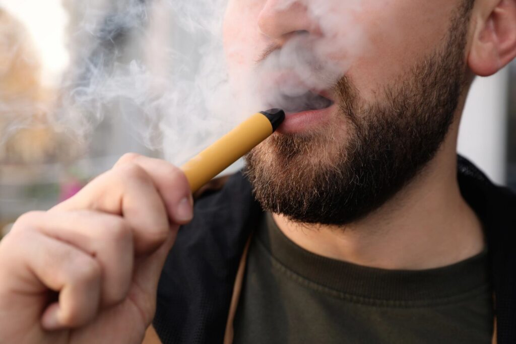 Close up of a man using a yellow disposable e-cigarettes Tulsa device while exhaling a thick cloud of white vapor.