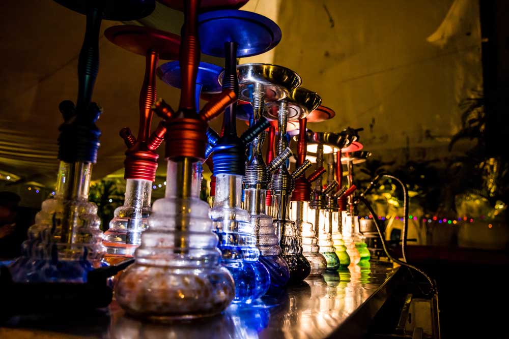 Row of glowing, modern hookahs with colorful glass bases and metallic stems in a dimly lit bar, a premium head shop Tulsa find.
